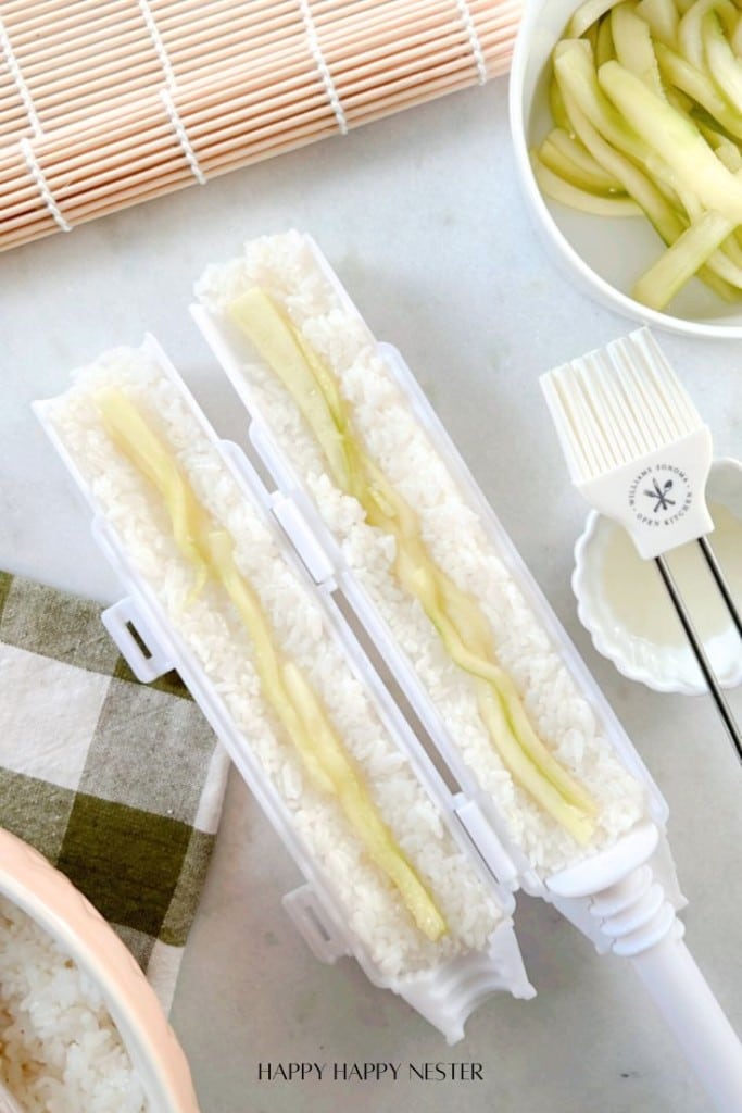 A sushi bazooka filled with rice and cucumbers is shown open, ready to be rolled. Nearby are a bowl of sliced cucumbers, a spatula, and a green checkered napkin, set on a light countertop with a sushi mat at the top.