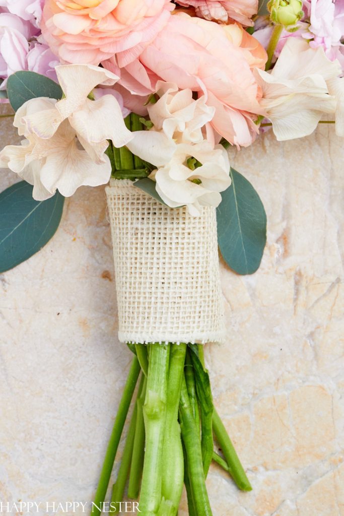 A close-up of a bouquet wrapped in burlap, featuring exquisite pink and peach flowers with green leaves, showcasing the charm of pink crafts against a light, textured stone background.