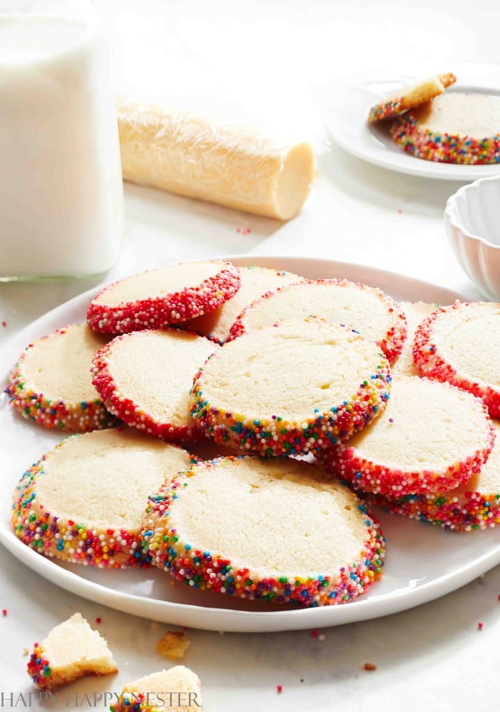 A plate of round cookies with edges coated in vibrant sprinkles, perfect examples of pink desserts, stands invitingly. In the background, a glass of milk accompanies more cookies on another plate, and a partially unwrapped roll of dough hints at more sweet creations to come.