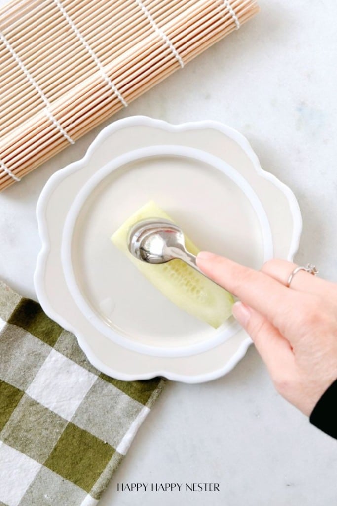 A hand holds a metal spoon, expertly scooping seeds from a cucumber slice on a white plate. Nearby, a Sushi Bazooka hints at creative culinary fun, while a checkered cloth and bamboo mat rest elegantly on the marble surface.