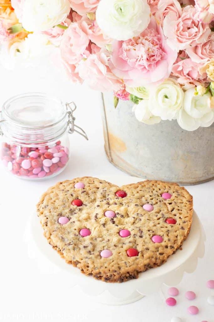 A heart-shaped cookie topped with pink and red candies sits on a white plate, embodying the essence of pink desserts. Beside it is a glass jar filled with matching sweets. In the background, a metal bucket overflows with pastel blooms, including delicate pink and white roses.