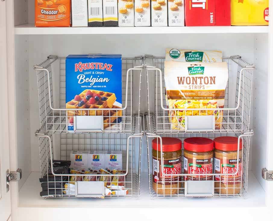 A pantry shelf, expertly organized with wire baskets, showcases professional organizing tips in action. The upper shelf holds boxes of Krusteaz Belgian waffle mix and wonton strips, while the lower shelf neatly displays jars of peanut butter and snack bars.