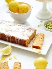 A healthy lemon loaf cake with a glaze sits on a white plate, nestled among sliced lemons and zest. Nearby, a white bowl overflows with whole lemons, while the background features a cake stand adorned with more lemons, accentuating this vibrant lemon loaf recipe.