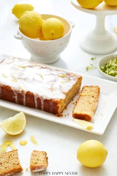 A healthy lemon loaf cake with a glaze sits on a white plate, nestled among sliced lemons and zest. Nearby, a white bowl overflows with whole lemons, while the background features a cake stand adorned with more lemons, accentuating this vibrant lemon loaf recipe.