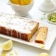 A healthy lemon loaf cake with a glaze sits on a white plate, nestled among sliced lemons and zest. Nearby, a white bowl overflows with whole lemons, while the background features a cake stand adorned with more lemons, accentuating this vibrant lemon loaf recipe.