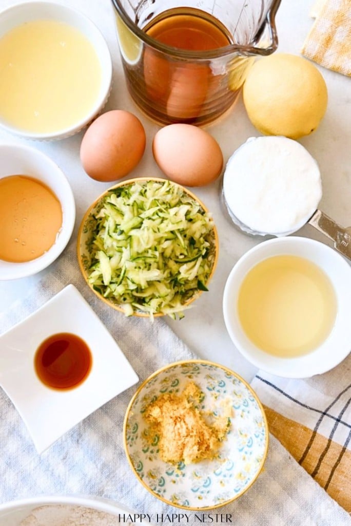 A table with ingredients for a healthy lemon loaf recipe: eggs, grated zucchini, lemon, milk, honey, vanilla extract, flour, brown sugar, and oil. They're all neatly arranged in bowls and measuring cups on a white surface with a checkered cloth.