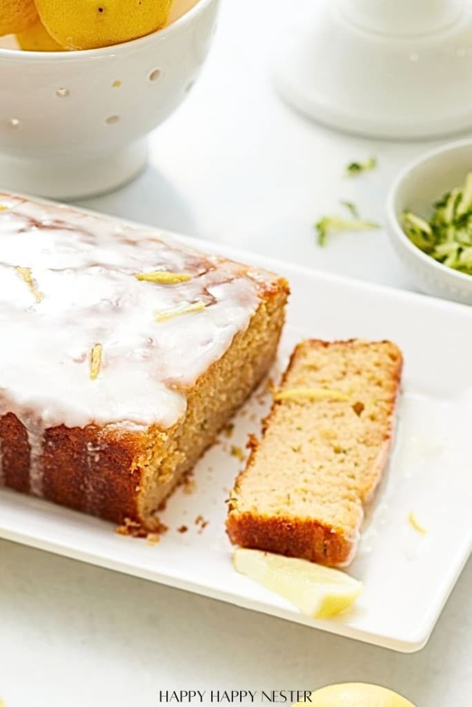 A glazed loaf of lemon zucchini bread on a white plate. One slice is cut and lay next to the loaf. A bowl of lemon slices and zucchini shreds are in the background.