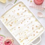 A rectangular white dish cradles the Recipe for Lemon Lush Dessert, topped with crushed nuts. Surrounding the dish are a pink glass, a spoon, lemon halves, tulips, a pink flower, and a pink checkered cloth.