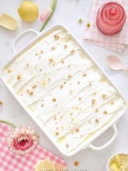 A rectangular white dish cradles the Recipe for Lemon Lush Dessert, topped with crushed nuts. Surrounding the dish are a pink glass, a spoon, lemon halves, tulips, a pink flower, and a pink checkered cloth.