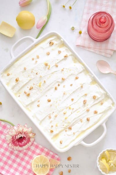 A rectangular white dish cradles the Recipe for Lemon Lush Dessert, topped with crushed nuts. Surrounding the dish are a pink glass, a spoon, lemon halves, tulips, a pink flower, and a pink checkered cloth.