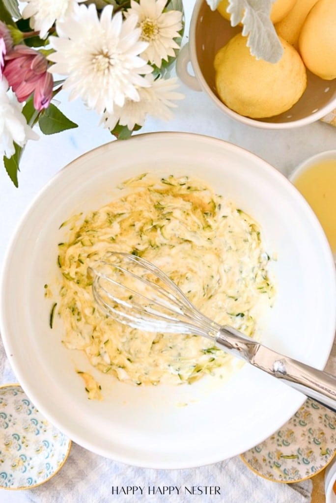 A white bowl filled with a creamy mixture and a whisk on top of a light tablecloth. Next to it are flowers and a bowl of lemons. The scene is bright and inviting.