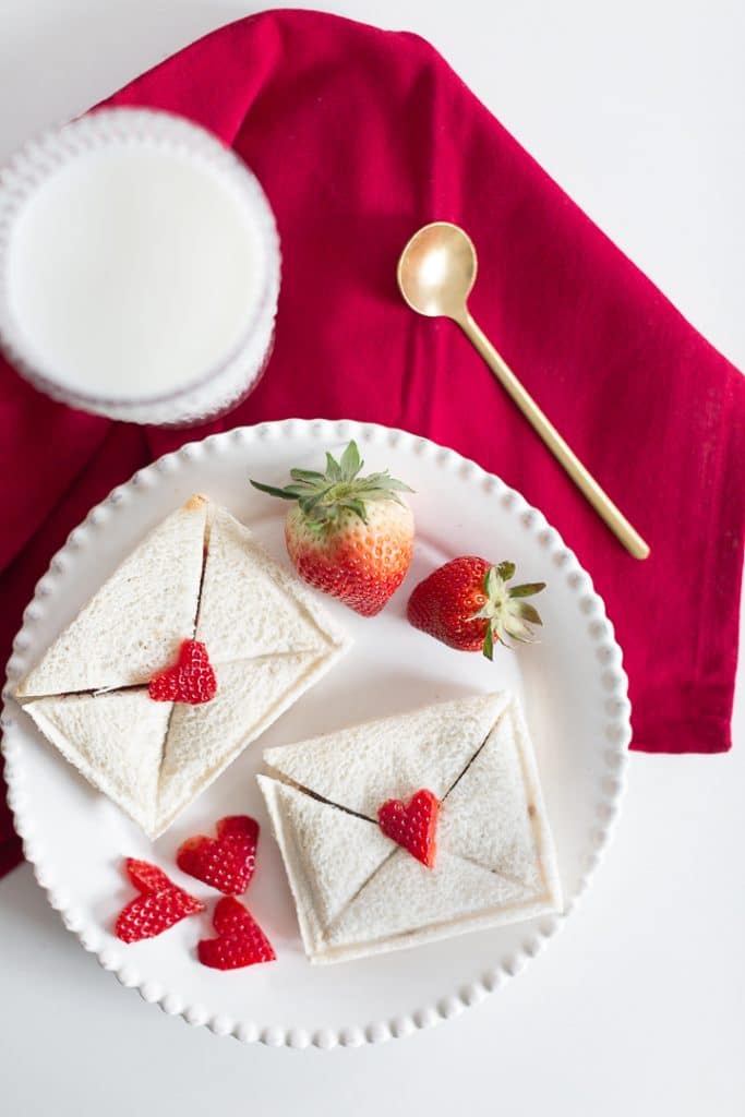 A plate with two sandwich halves shaped like envelopes, each adorned with a strawberry heart, resembles an easy fruit trifle recipe. Additional heart-shaped strawberry slices and whole strawberries are scattered around. A glass of milk and a spoon rest on a red cloth nearby.