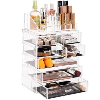 Clear acrylic makeup organizer with multiple drawers and compartments, designed using professional organizing tips. It holds lipsticks, foundations, brushes, a fragrance bottle, and more. Various makeup products are neatly displayed on top and inside the transparent drawers.