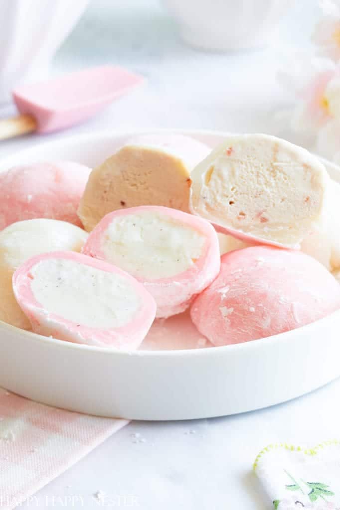 A white plate holds several pieces of pink desserts, featuring mochi ice cream with pink and white exteriors. Some are cut open to reveal creamy fillings. A light pink spatula and a flower are faintly visible in the background.