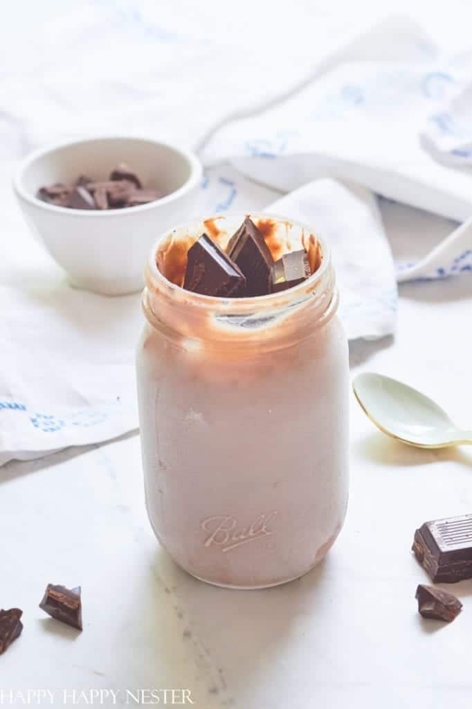 A glass jar filled with chocolate ice cream topped with rich chocolate chunks sits on a white surface. Nearby, a spoon and a small bowl overflowing with chocolate pieces hint at delicious recipes made with chocolate chips, alongside an elegant white cloth.