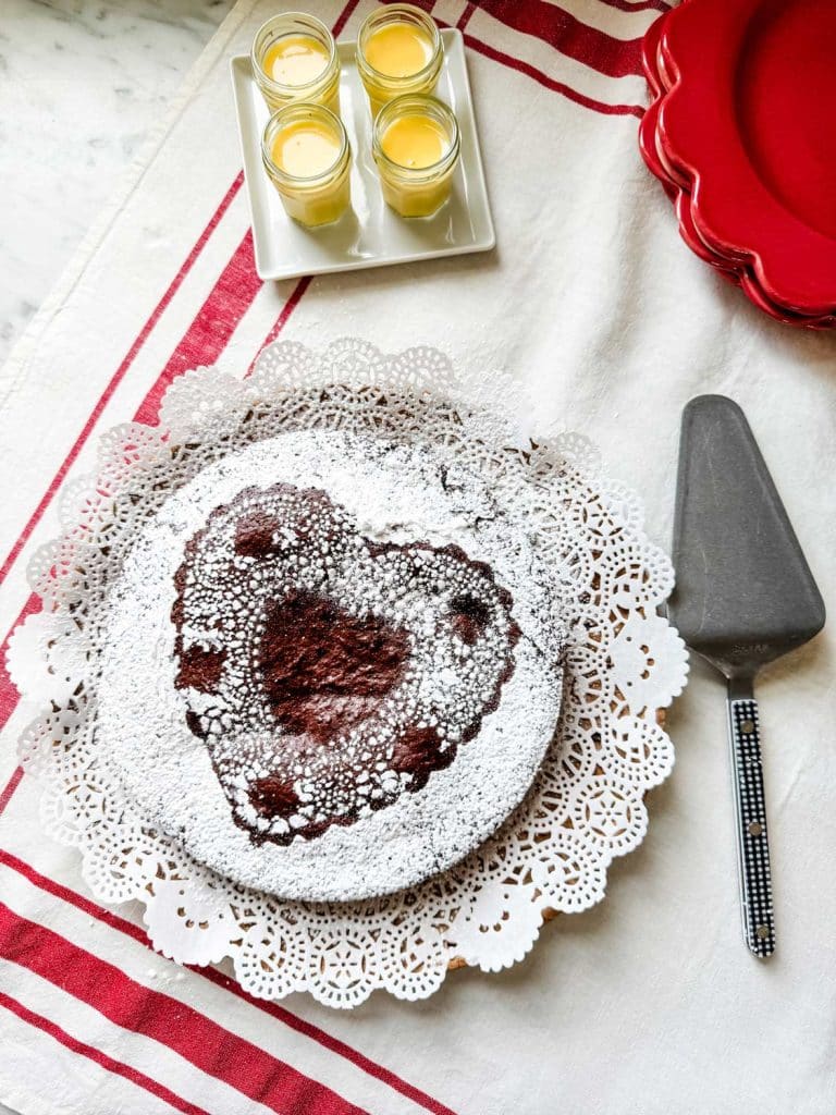 Heart-shaped chocolate cake dusted with powdered sugar on a lace doily. Next to it, a pie server and glasses of yellow liquid sit on a square plate atop a red and white striped tablecloth, making this scene as delightful as an easy fruit trifle recipe.