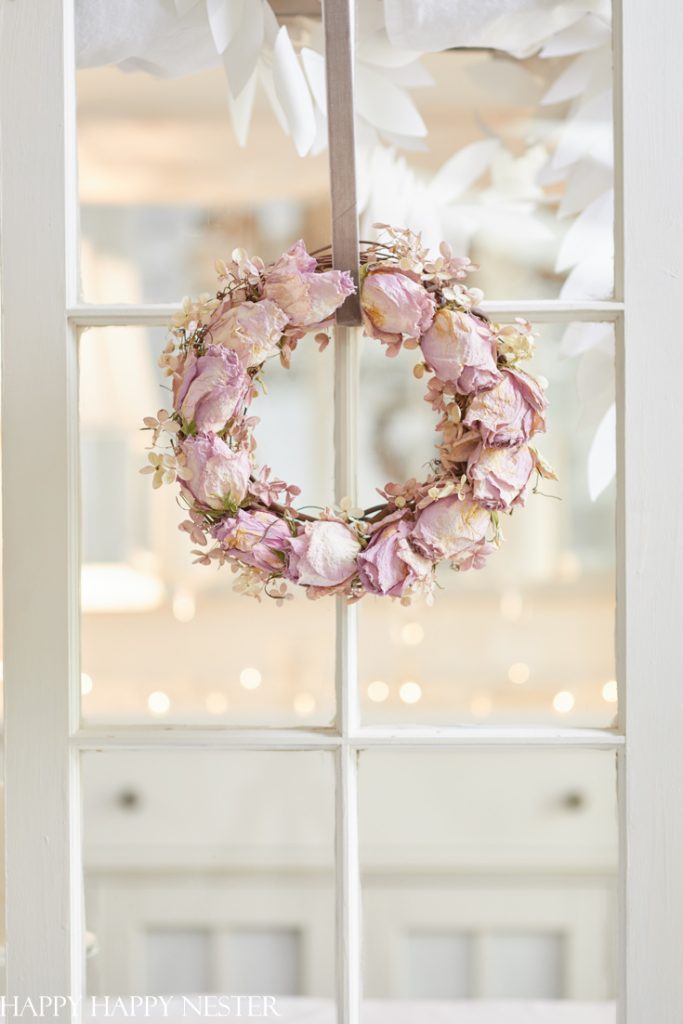 A delicate pink and white floral wreath, a masterpiece of pink crafts, hangs on a glass-paned door. The background is softly blurred, creating a dreamy, light-filled atmosphere.