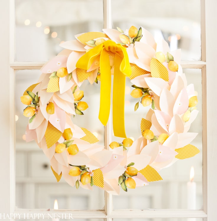 A decorative wreath made of paper leaves and lemons, accented with a large yellow ribbon, hangs on a glass door. This charming seasonal wreath features a mix of soft pink and yellow colors, evoking a cheerful and fresh appearance.