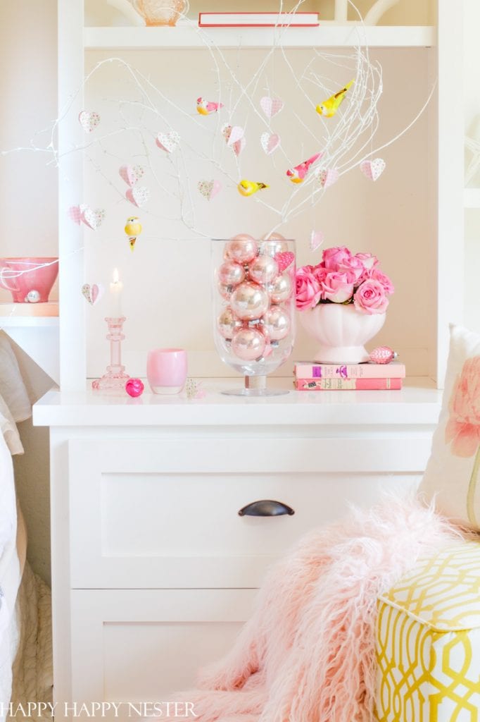 A cozy, pastel-themed interior features a white cabinet displaying pink crafts and ornaments in a glass vase, roses in a white pot, and books. Soft decorative items, like a fluffy pink blanket, add to the serene ambiance.