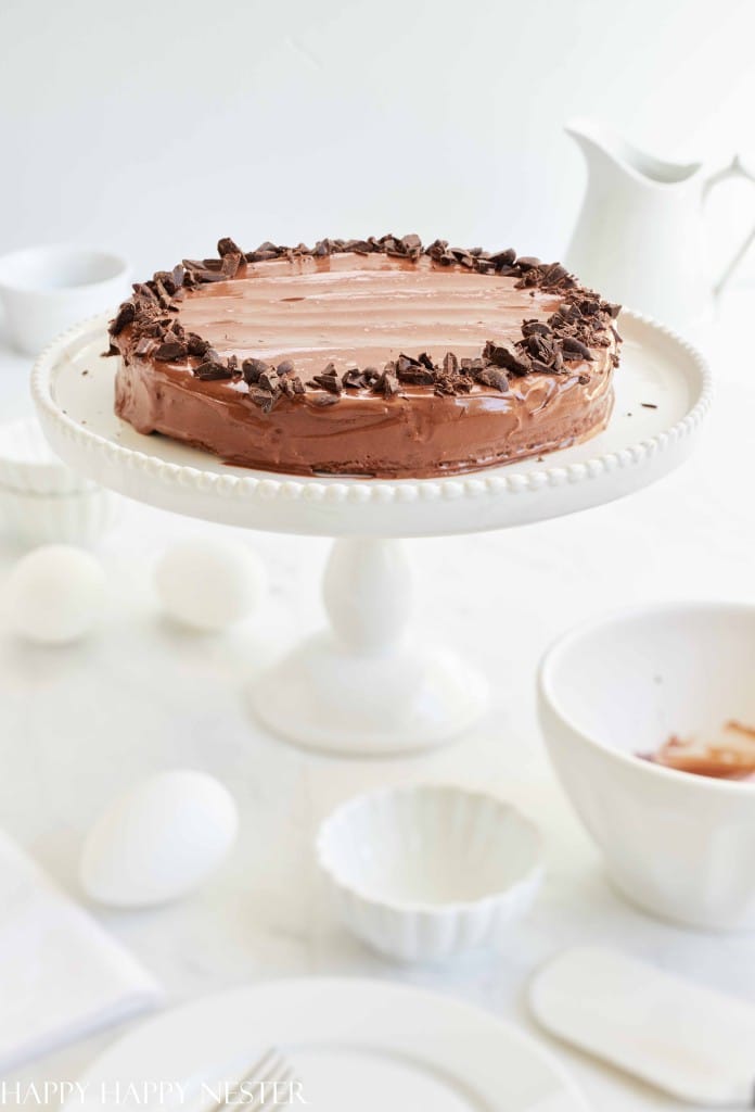 A chocolate cake with a smooth frosting, enriched by a scattering of chocolate shavings and chips, sits elegantly on a white cake stand. The backdrop features white bowls, eggs, and a milk jug on a pristine surface, perfect for those who savor recipes made with chocolate chips.