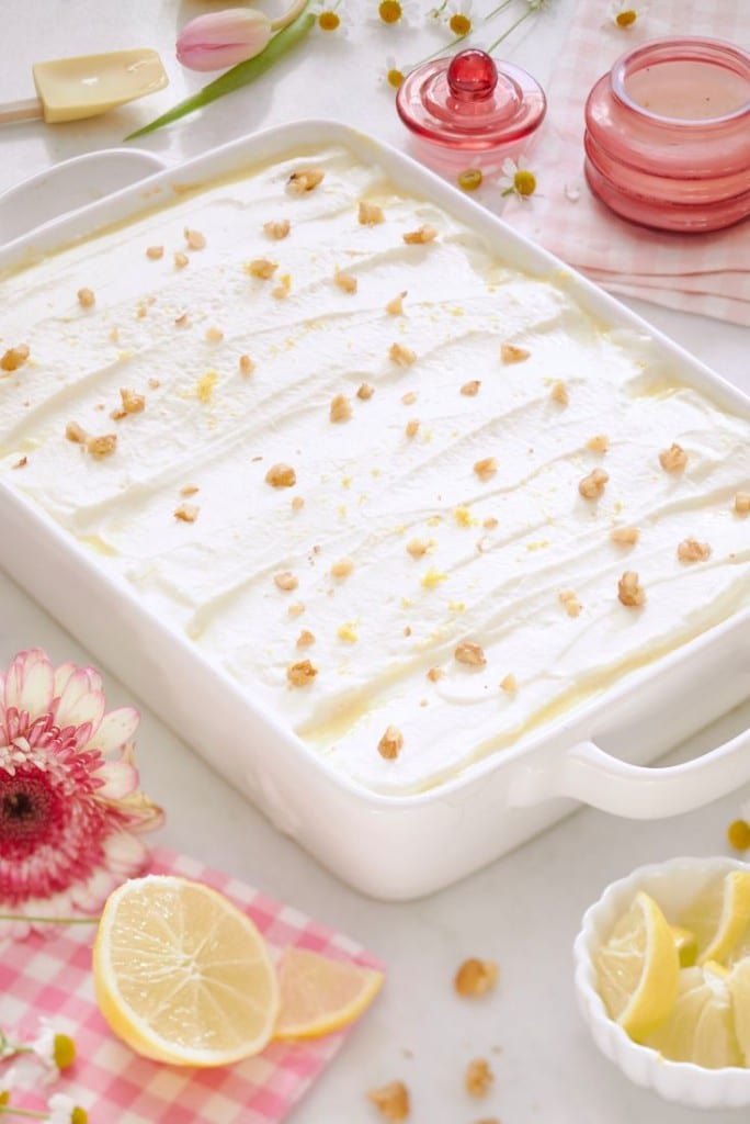 A white rectangular dish filled with whipped cream topped with chopped nuts, surrounded by pink flowers, a lemon wedge, sliced lemon in a bowl, and decorative pink elements on a checkered cloth.