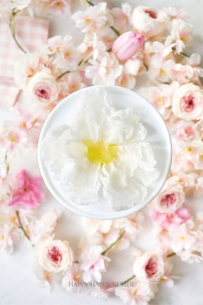 A delicate white flower with a yellow center rests in a white dish, set against a floral background of pink and white blossoms, reminiscent of artfully crafted fried rice paper flowers.