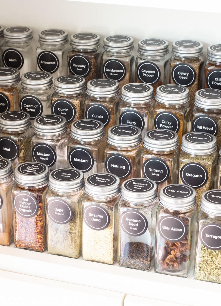 A neatly organized spice rack showcases clear jars labeled with spices like cayenne pepper, curry, oregano, mustard, and nutmeg. With professional organizing tips in play, the jars are arranged in precise rows on a white shelf.