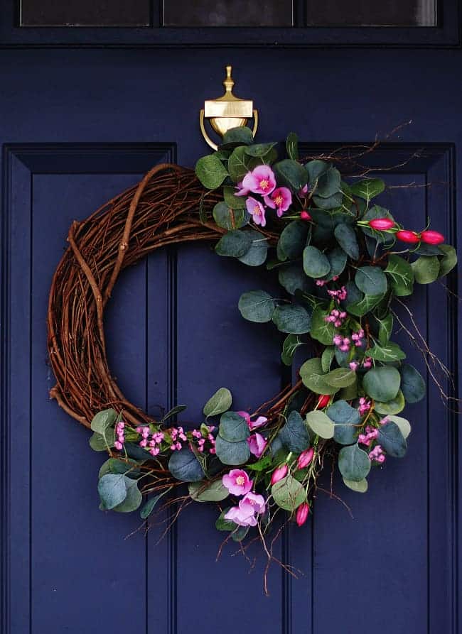 A dark blue door with a gold knocker is adorned with a seasonal wreath made of twisted branches, green eucalyptus leaves, and pink flowers like orchids and tulips, arranged asymmetrically on one side.