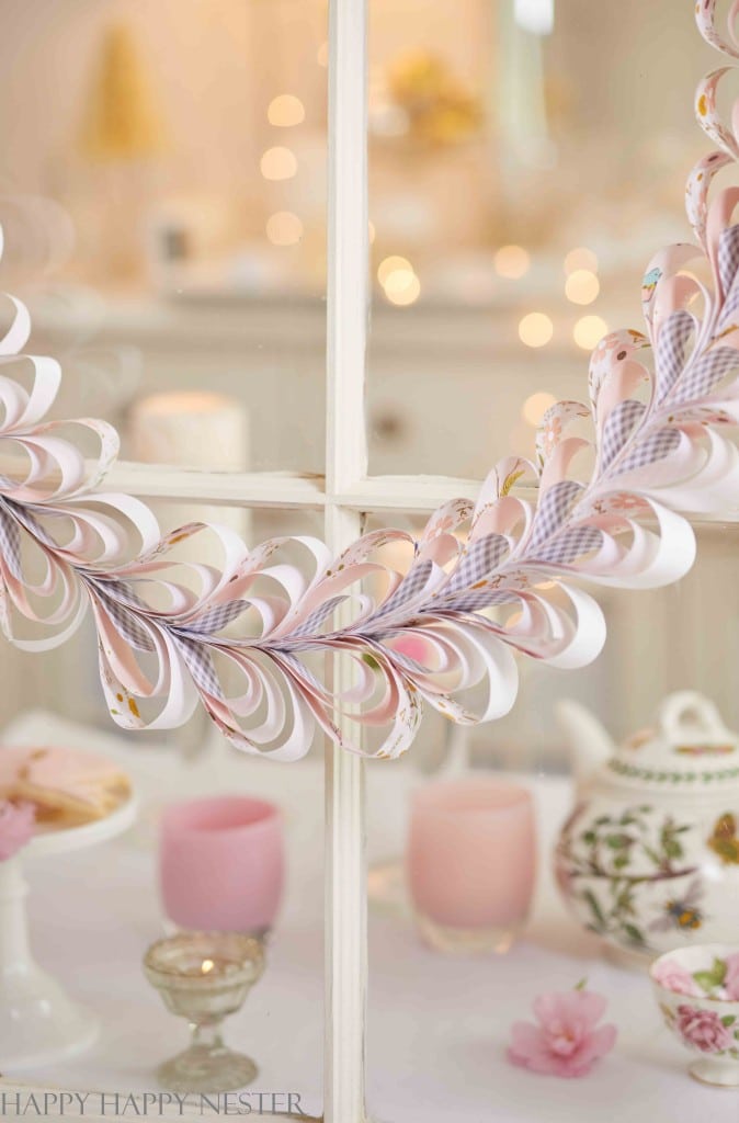 A delicate paper garland featuring heart and teardrop shapes hangs on a window with a blurred, softly lit background. Below are a teapot with floral patterns, pink candles, and small decorative items crafted in soothing shades of pink on a white surface.