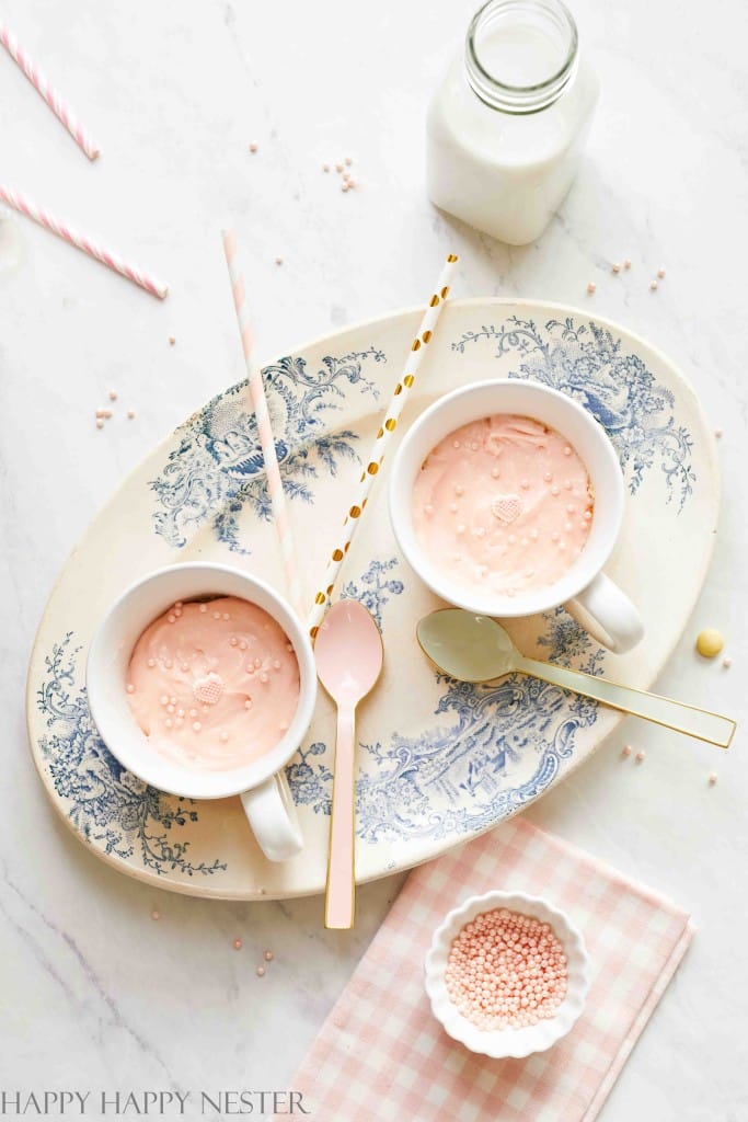 Two cups of pink milk with gold and pink straws sit on an oval tray with a blue floral pattern. Beside them, a gold and a pink spoon rest alongside the tempting scene of pink desserts. A small bowl filled with pink sprinkles and a bottle of milk complete the setup on a pink gingham cloth.