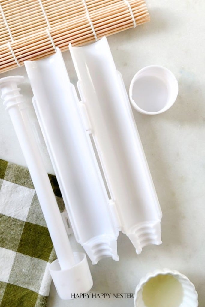 Top view of sushi-making tools on a light surface. Includes a white cylindrical sushi mold, a bamboo rolling mat, and a small white container. A green and white checkered cloth is partially visible in the corner.