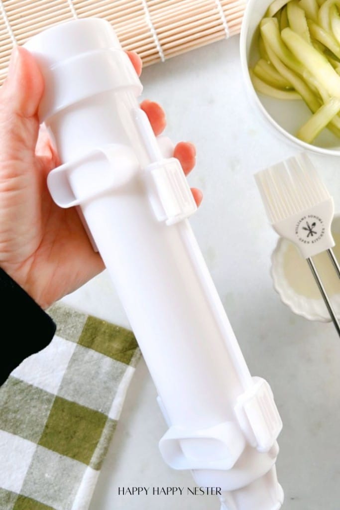 A hand holding a white plastic sushi roller next to a bamboo mat, sliced cucumbers in a bowl, a pastry brush, and a green checkered cloth on a marble surface.