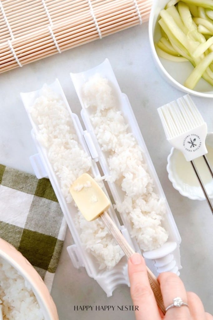 A person uses a spatula to press sushi rice into a white mold, reminiscent of a sushi bazooka. Nearby, a bowl holds sliced cucumbers, and a bamboo sushi mat peeks out. A cloth with a green and white checkered pattern lies on the side.