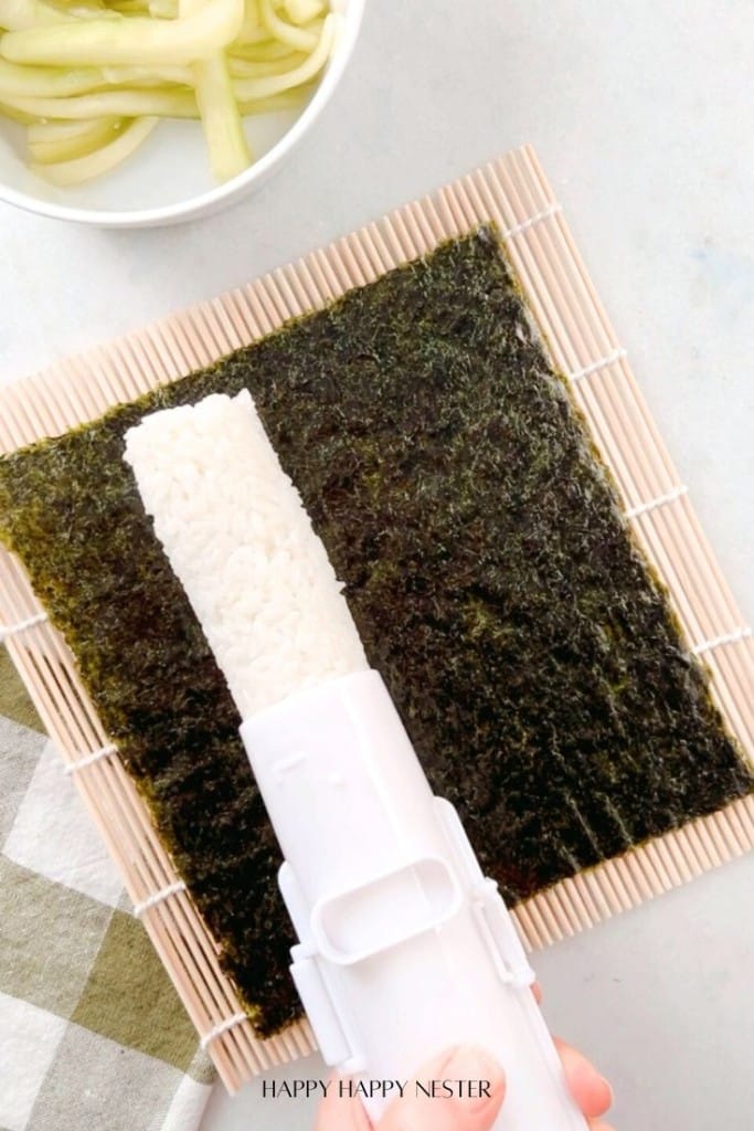 A bamboo sushi mat with a sheet of nori and rice being spread on it using a white sushi rolling tool. A bowl containing cucumber strips is in the background.