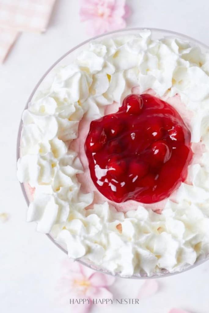 A dessert with a red fruit topping, possibly cherry, in the center, surrounded by swirls of white whipped cream. The background is blurred with soft pink hues and abstract elements.