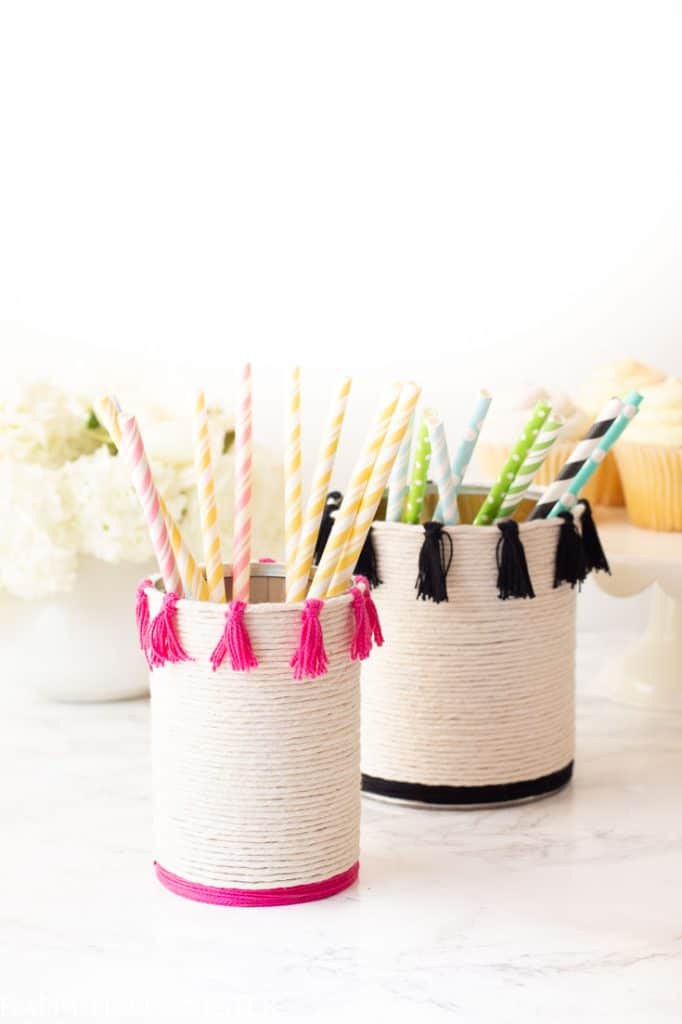 Two cylindrical containers wrapped in white twine, one adorned with pink crafts like tassels and the other with black, hold colorful straws. The background features white flowers and cupcakes resting on a marble surface.