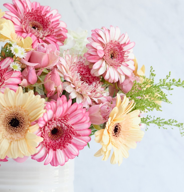 A bouquet in a white vase showcases pink and peach gerbera daisies and roses, surrounded by green foliage and small white flowers. Its delicate arrangement is perfect for adding a touch of elegance to any space or inspiring pink crafts against a light background.