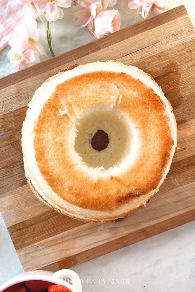 A toasted bagel with a golden brown top sits on a wooden cutting board, evoking the simple pleasures of an easy morning. Nearby, pink flowers and a bowl of sliced strawberries bring to mind the vibrant ingredients of an easy fruit trifle recipe.