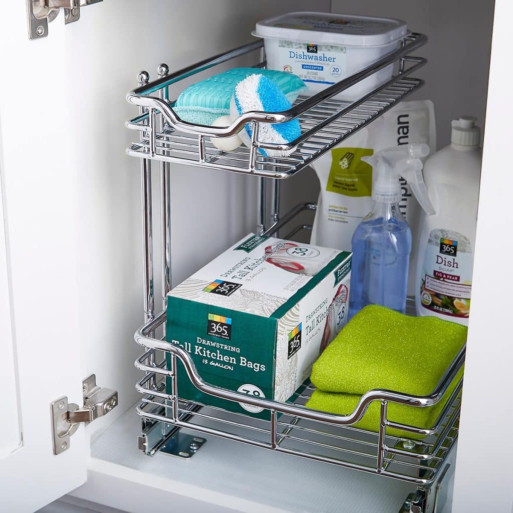 A kitchen cabinet with a chrome pull-out organizer, expertly designed with professional organizing tips. It holds dishwasher detergent, a box of kitchen bags, cleaning sprays, sponges, and a green cloth. The two-level organizer ensures your cleaning supplies are neatly stored and easily accessible.