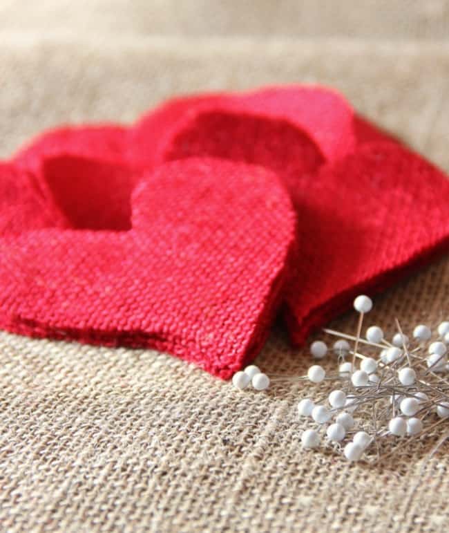 Red fabric hearts stacked on a burlap surface create a charming setting for pink crafts, with a cluster of white-headed pins nearby adding a touch of whimsy.