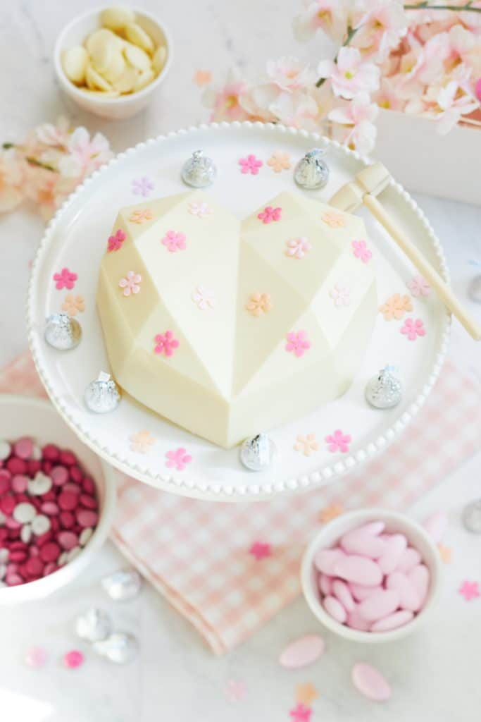 A heart-shaped white chocolate cake with pink and peach floral decorations sits elegantly on a white stand. This centerpiece is accompanied by an array of pink desserts, including chocolate kisses, bowls of pink candies, and blossoms scattered across a pastel checked cloth.