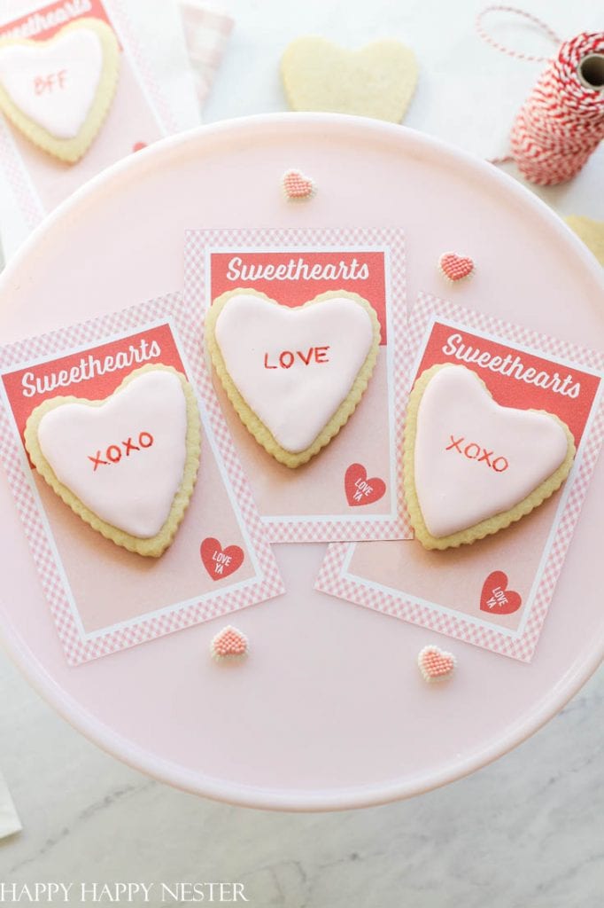 Heart-shaped cookies with pink icing rest on cards labeled "Sweethearts," featuring "XOXO" and "LOVE." Nearby, small heart sprinkles and a spool of red twine add to the charm of this pink crafts display on a vibrant platter.