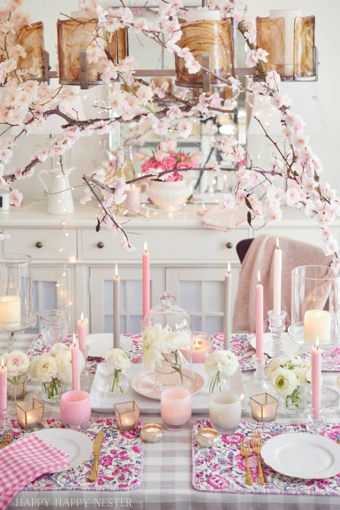 A beautifully set dining table with pink and white floral placemats showcases pink crafts, alongside various candles and white flowers in glass vases. Overhead, branches adorned with pink blossoms complete the soft and elegant ambiance.