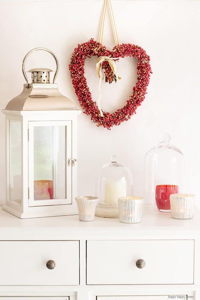 A white dresser features a silver lantern, three glass cloches covering candles, and two uncovered candles. Adding to the cozy elegance, a heart-shaped wreath of red berries hangs above, with delicate touches of pink crafts enhancing the charming display.