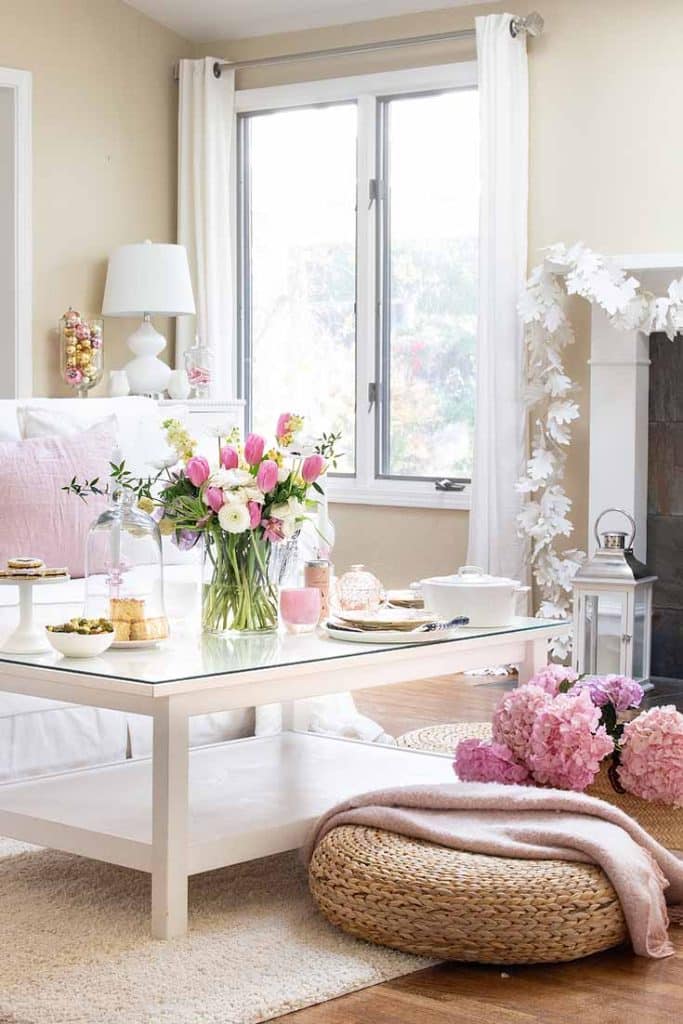 Bright living room with a white coffee table showcasing a vase of pink and white flowers, ideal for Valentine's Day decor. Decorative items complement the scene while a natural woven pouf sits on a light rug next to pink hydrangeas. White curtains frame the window, enhancing the airy feel.