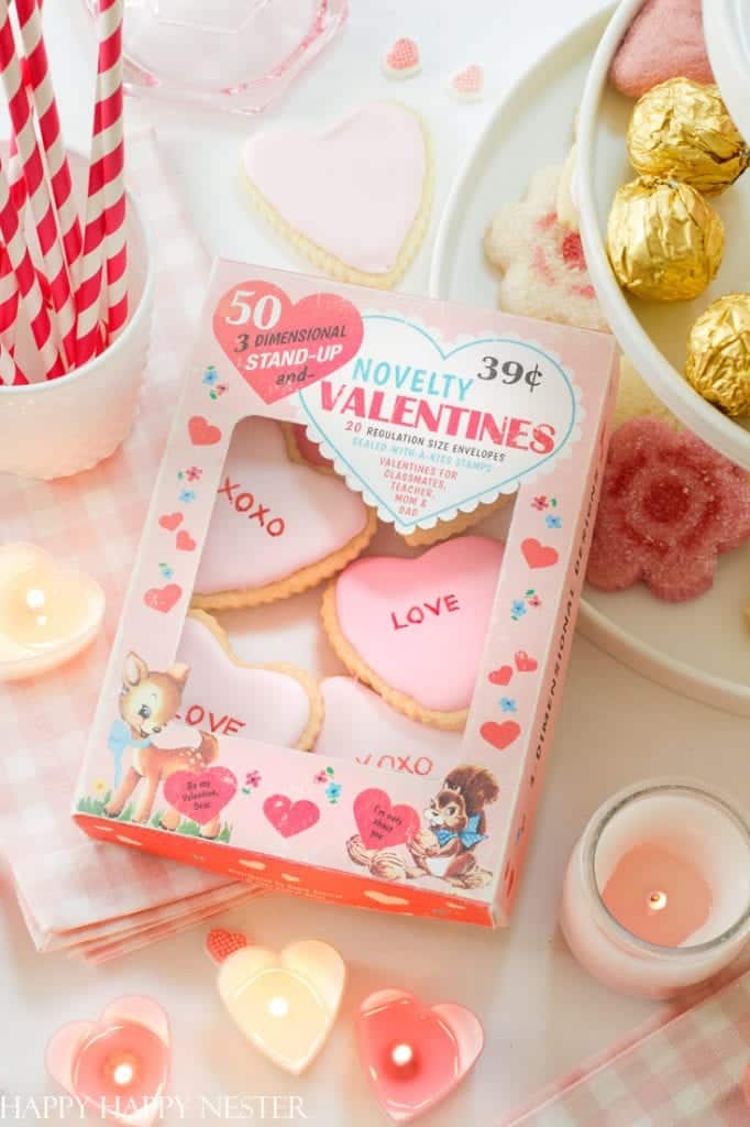 A vintage-style box of novelty valentines features pink and white heart-shaped cookies labeled "XOXO" and "LOVE," surrounded by delicate pink crafts, red striped straws, and gold-wrapped chocolates on a light checkered tablecloth.