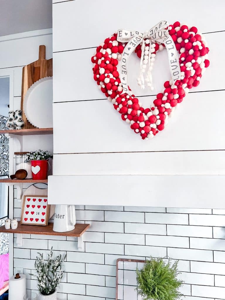 A decorative kitchen scene features a heart-shaped red and white pom-pom wreath hanging on a white paneled wall, highlighting the charm of seasonal wreaths. It's adorned with a ribbon reading "love." Nearby, shelves display dishes and decor, including a jug labeled "water.