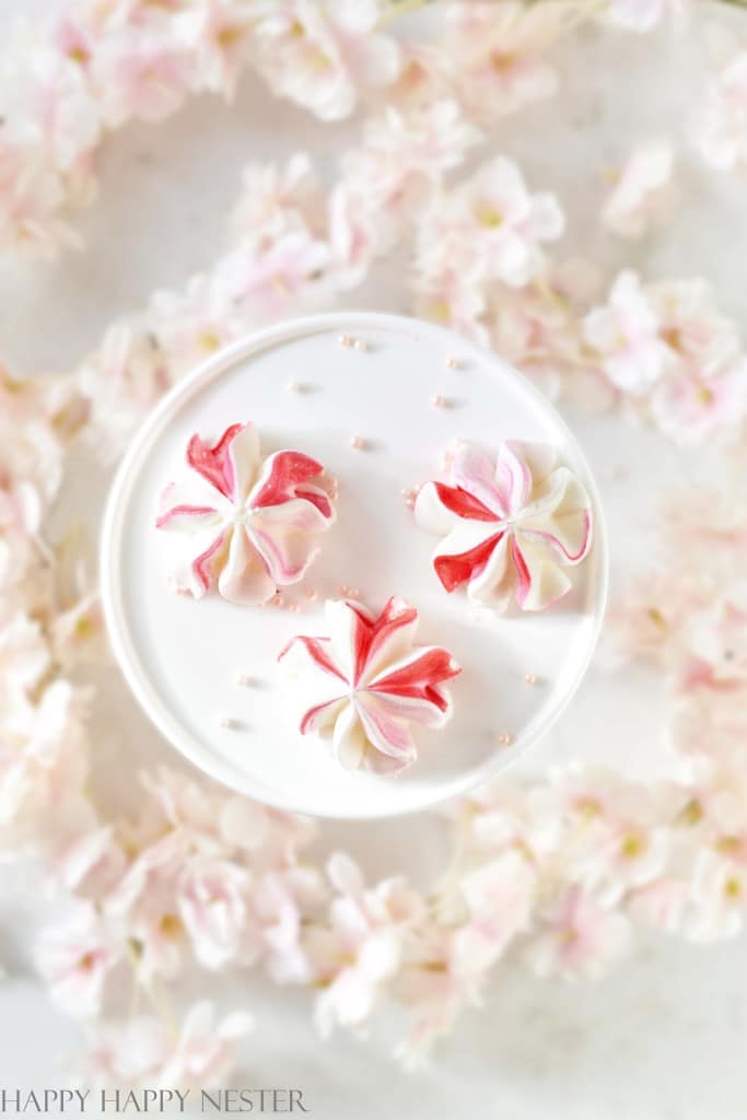 Three pink and white meringue cookies, perfect pink desserts, sit on a round white plate surrounded by soft pink cherry blossoms. The cookies boast a delicate swirled pattern, with light sprinkles scattered artistically across the plate.