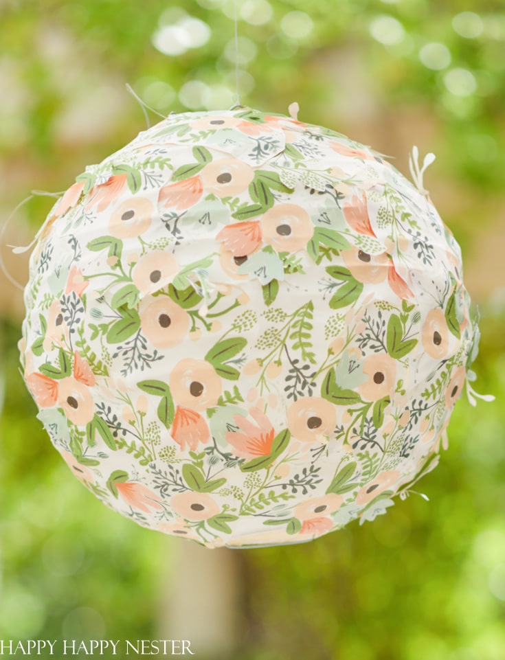 A round hanging lantern, crafted as an easy flower paper project, is adorned with pastel hues and floral designs featuring pink and peach blossoms with green leaves. It elegantly stands out against a softly blurred backdrop of greenery.