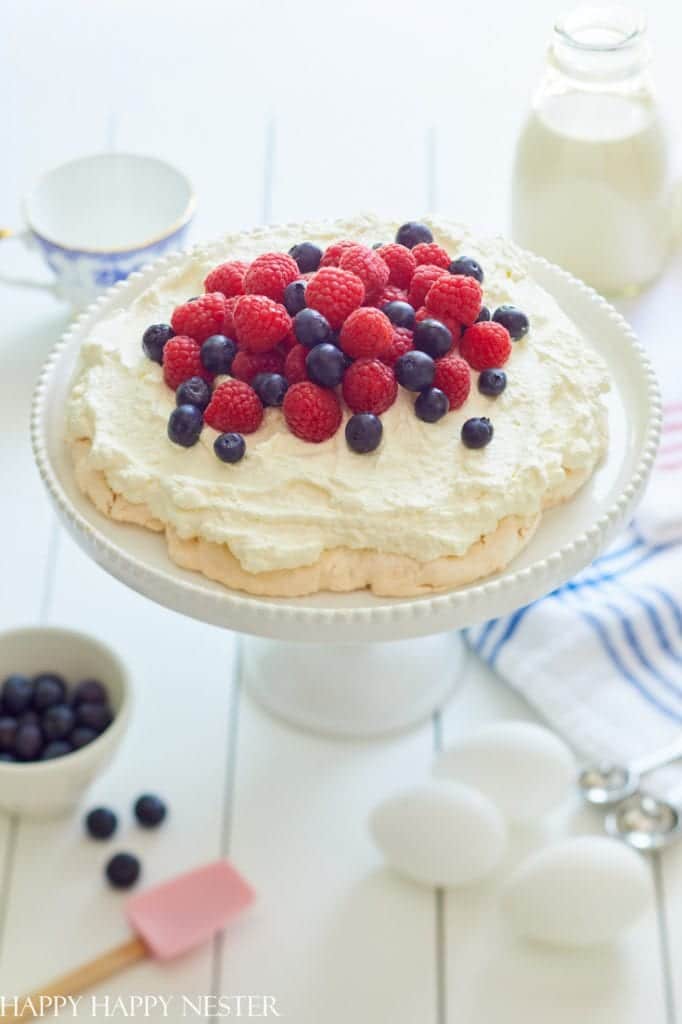 A pavlova, one of my favorite desserts, is topped with fresh raspberries and blueberries on a white cake stand. Around it are a small bowl of blueberries, eggs, a milk bottle, a cup, a pink spatula, and a striped cloth on the rustic white wooden table.